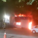 Armazém pegando fogo, carros perto e um bombeiros indo combater o incêndio.