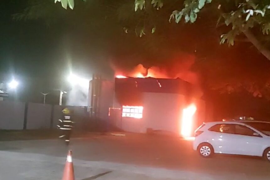 Armazém pegando fogo, carros perto e um bombeiros indo combater o incêndio.